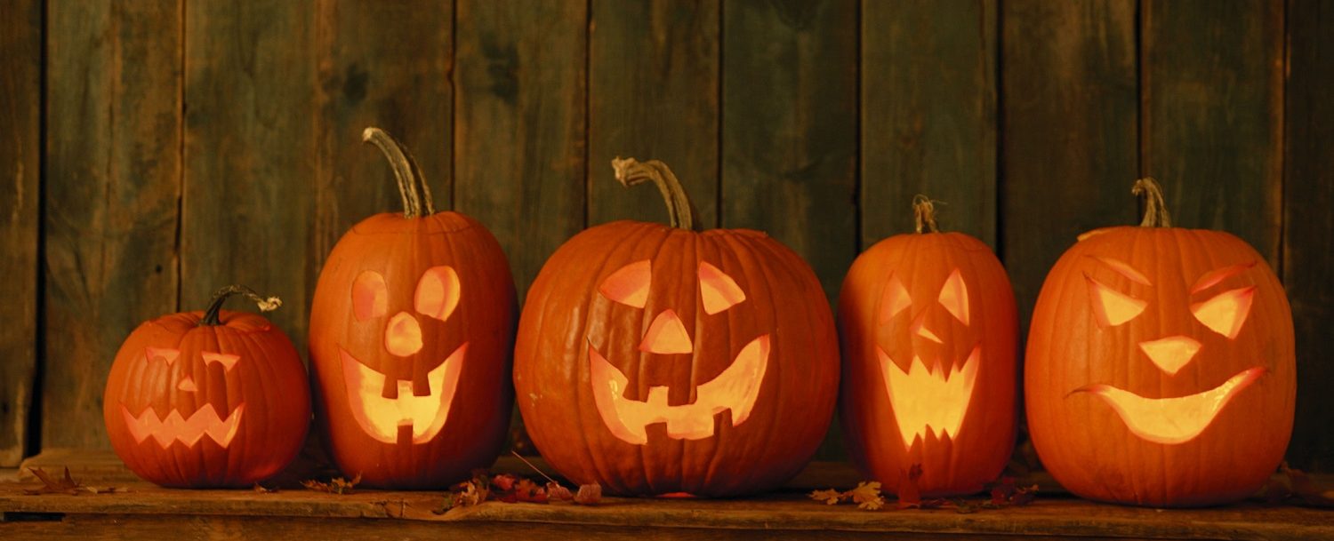 Halloween picture showing five pumpkins that have been carved. They are lite up with candles with a wood background.