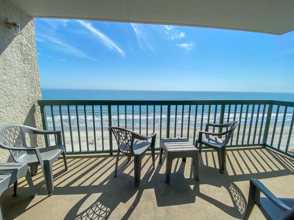 an outdoor balcony with the blue sky with the ocean and beach in view along with three small chairs and a small table on the balcony