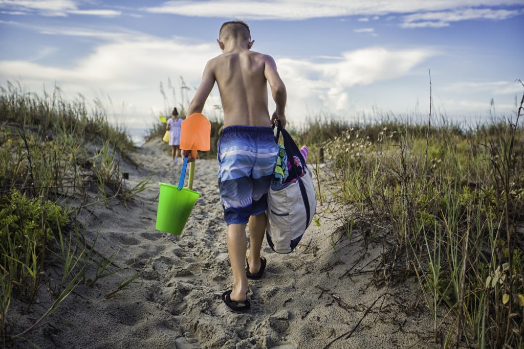 young boy carrying a tote bag with towels and shovel and beach bucket along with a young girl on the way to the beach for the day