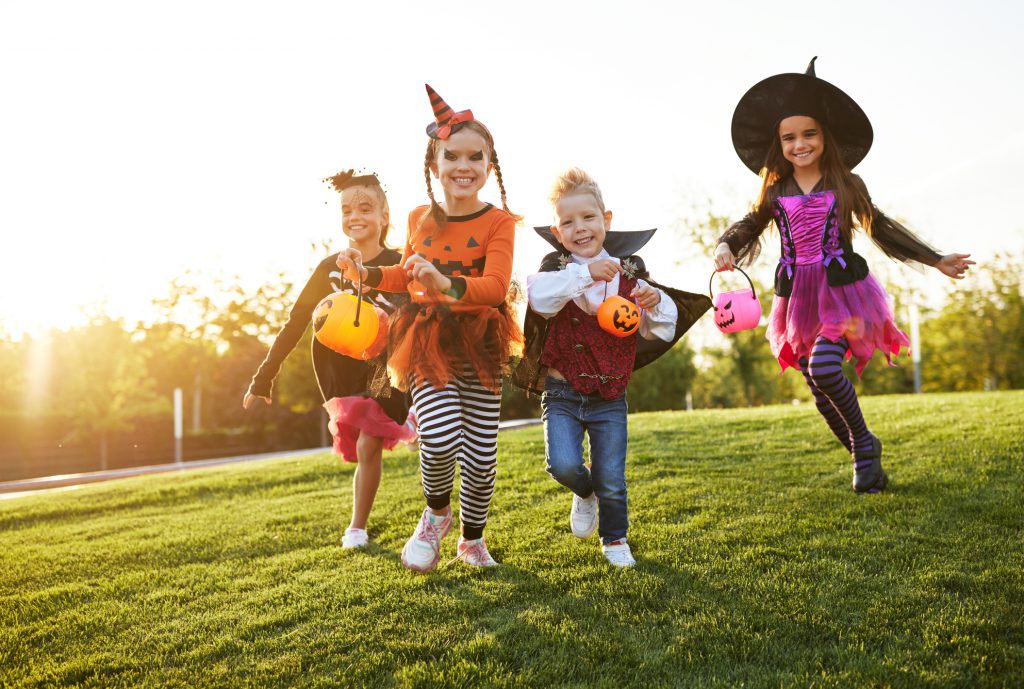 Group of excited kids in spooky costumes smiling and running on lawn during Halloween celebration in evening in park