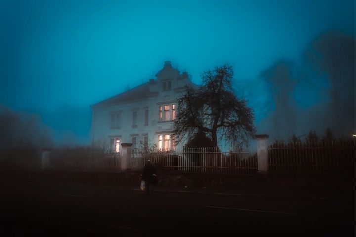 picture of a haunted house with fog around at dusk with lights on. 